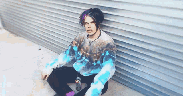 a young man with blue and pink hair is sitting on the ground with a bottle of soda .