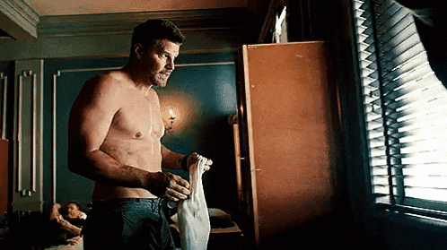 a shirtless man is standing in front of a window in a bedroom holding a towel .