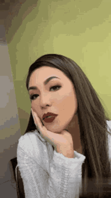 a woman wearing red lipstick and a white sweater is taking a selfie with her hand on her face .