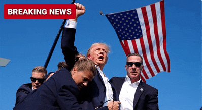 a man in a suit holds up his fist in front of an american flag with breaking news written on the bottom