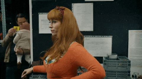 a woman in an orange sweater is standing in front of a bulletin board with information on it