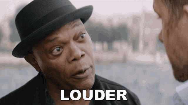 a man wearing a hat is talking to another man and the word louder is on the screen in front of him