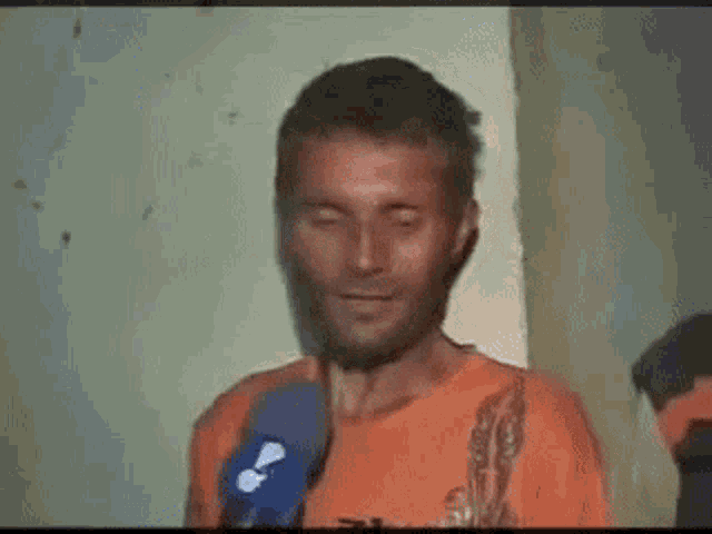 a man in an orange shirt is talking into a microphone and smiling .