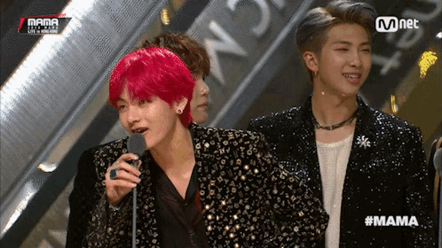 a man with red hair is speaking into a microphone while another man looks on