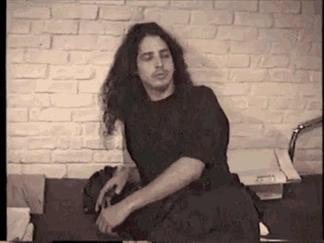 a man with long hair and a mustache is sitting in front of a brick wall in a room .