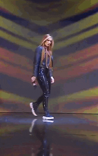 a woman wearing a leather jacket and black pants is walking on a stage .
