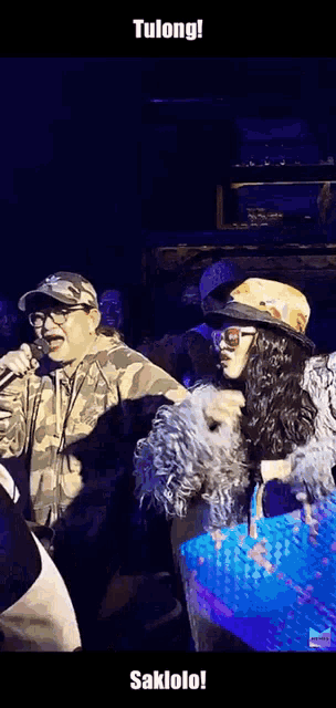 a man singing into a microphone next to a woman wearing sunglasses and a hat with the words tulong on it