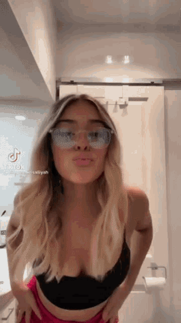 a woman wearing sunglasses and a black bra is standing in a bathroom .