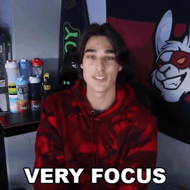 a young man in a red hoodie is sitting in front of a wall with a cat on it that says very focus