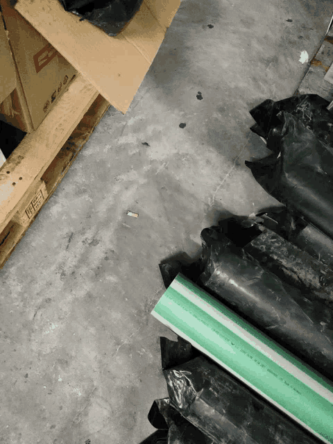 green and black pipes are stacked on a concrete floor next to a cardboard box that says ' e ' on it