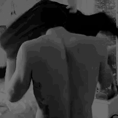 a black and white photo of a shirtless man taking off his shirt in a bedroom .