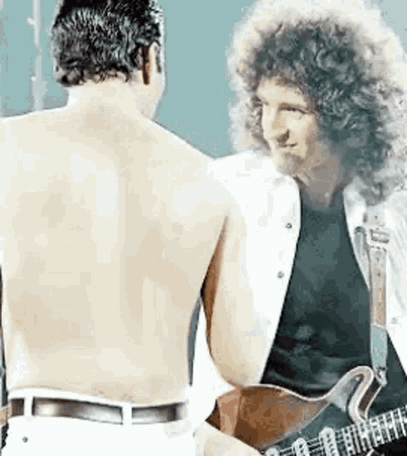 a man without a shirt is playing a guitar next to a man with a guitar .