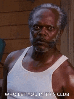 a man with a beard and a white tank top is standing in front of a building and making a funny face .