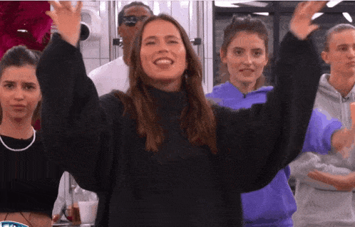 a woman in a black sweater stands in front of a group of people with their arms in the air
