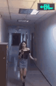 a woman is walking down a hallway with a green exit sign above her head
