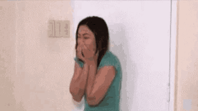 a woman in a green shirt is standing in front of a white wall covering her mouth .
