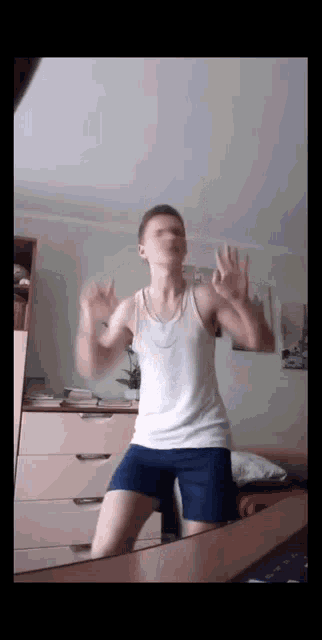 a man in a white tank top and blue shorts is dancing in front of a mirror in a bedroom .