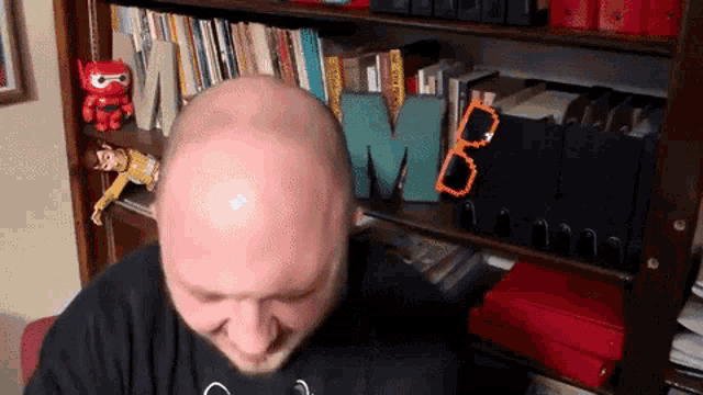 a bald man is sitting in front of a bookshelf with the letter m on it