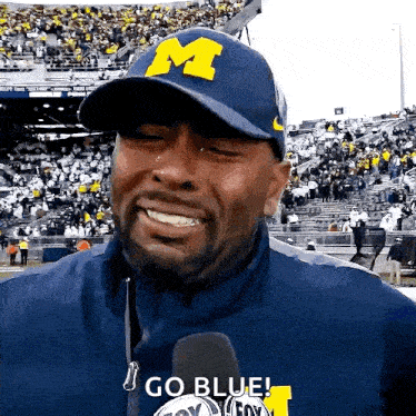 a man wearing a blue hat with the letter m on it is smiling and talking into a microphone .