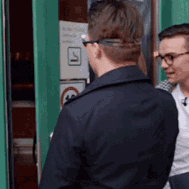 a man wearing glasses and a black jacket is standing in front of a green door .