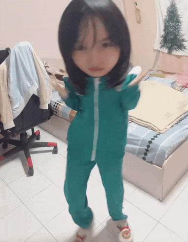 a little girl wearing a green jumpsuit is standing in a bedroom