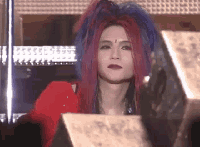 a woman with red and blue hair and red lipstick is sitting in a dark room .