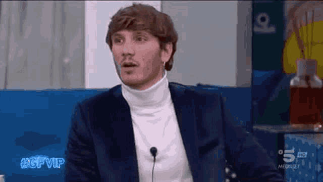 a man in a suit and turtleneck is sitting in front of a microphone on a television show .
