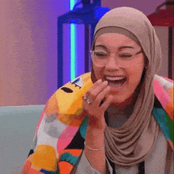 a woman in a hijab and glasses is laughing with her mouth open .