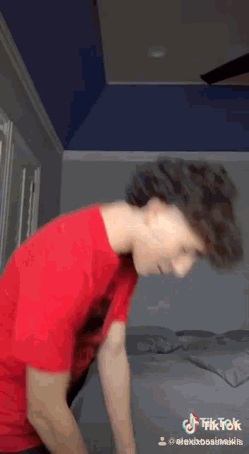 a young man in a red shirt is standing on a bed in a bedroom .