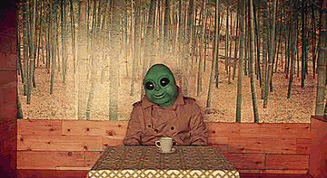 a man wearing a green alien mask is sitting at a table with a cup of coffee .