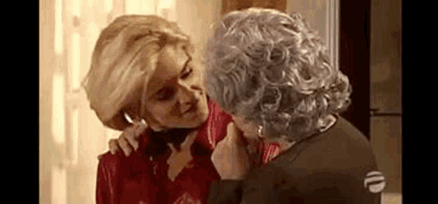 two women are hugging each other in a living room . one of the women is wearing a wig .