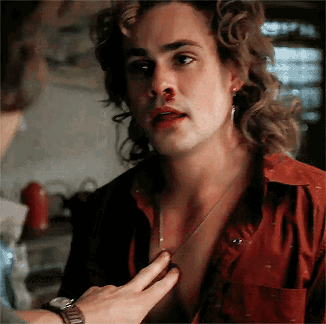 a man with long hair is being touched by another man 's hand while wearing a red shirt .