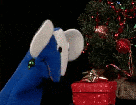 a blue and white mouse puppet is standing in front of a christmas tree and a gift box