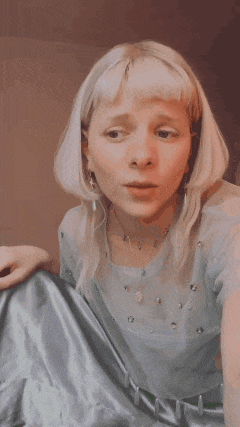 a woman with blonde hair is sitting on the floor wearing a blue shirt and a necklace .
