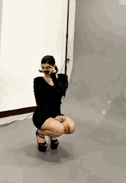 a woman in a black dress and heels is squatting down with her legs crossed