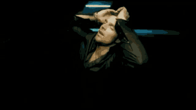 a man in a black shirt is sitting in a dark room with his hands behind his head .