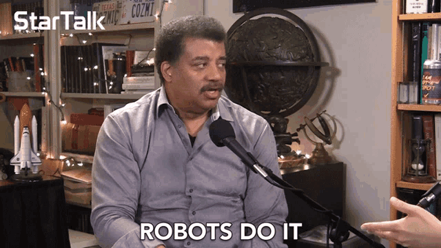 a man talking into a microphone with the words " robots do it " behind him