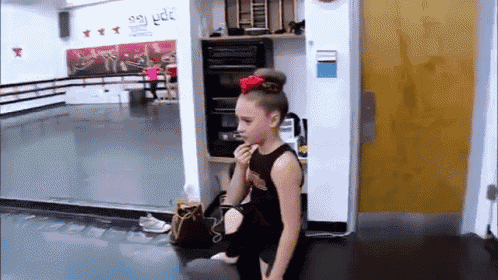 a girl with a bow in her hair is sitting in a dance studio eating a snack .