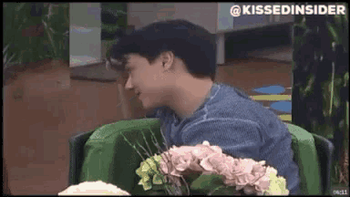 a man in a blue shirt is sitting in a green chair with flowers in front of him and the words kissedinsider on the bottom right