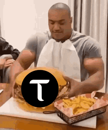 a man is sitting at a table eating a hamburger with the letter t on it