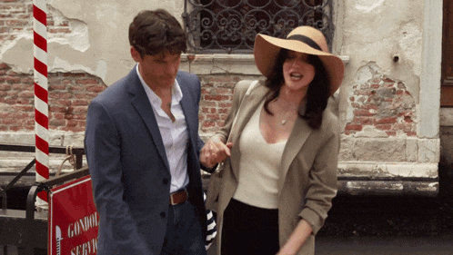 a man in a suit and a woman in a hat are walking in front of a sign that says gondola service