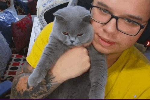 a man wearing glasses is holding a gray cat in his arms