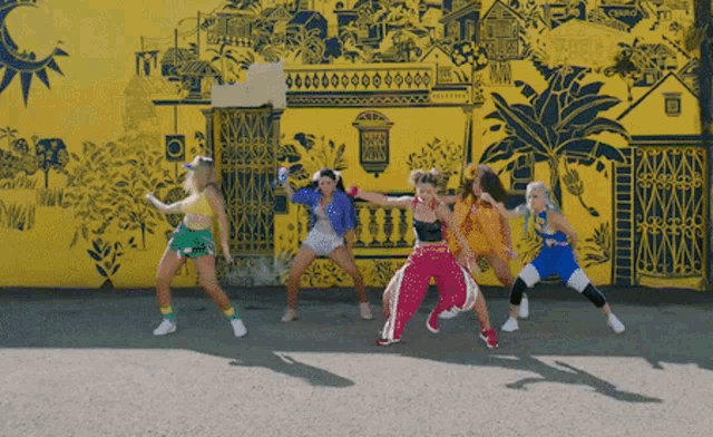 a group of young women are dancing in front of a yellow wall with a drawing of a building on it