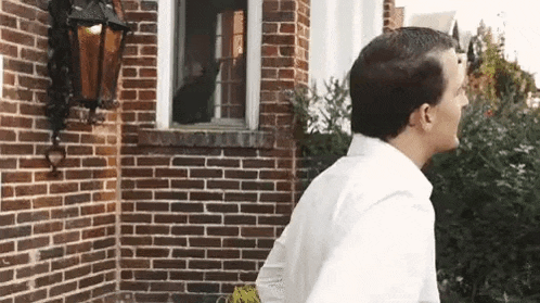 a man in a white shirt is standing in front of a brick building looking out a window .