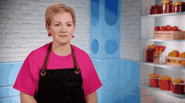 a woman in a pink shirt and black apron stands in front of a shelf full of jars
