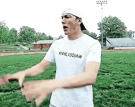 a man wearing a white hat and a white t-shirt is standing in a field with his hands outstretched .