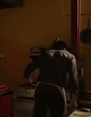 a man in a gray shirt and white apron stands in a kitchen