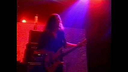 a man with long hair is playing a guitar in a dark room with purple lights behind him .