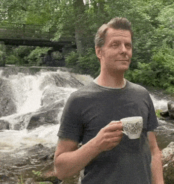 a man is standing next to a waterfall holding a cup of coffee .
