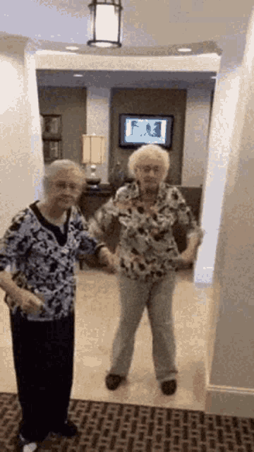 two older women are standing next to each other in a hallway holding hands .